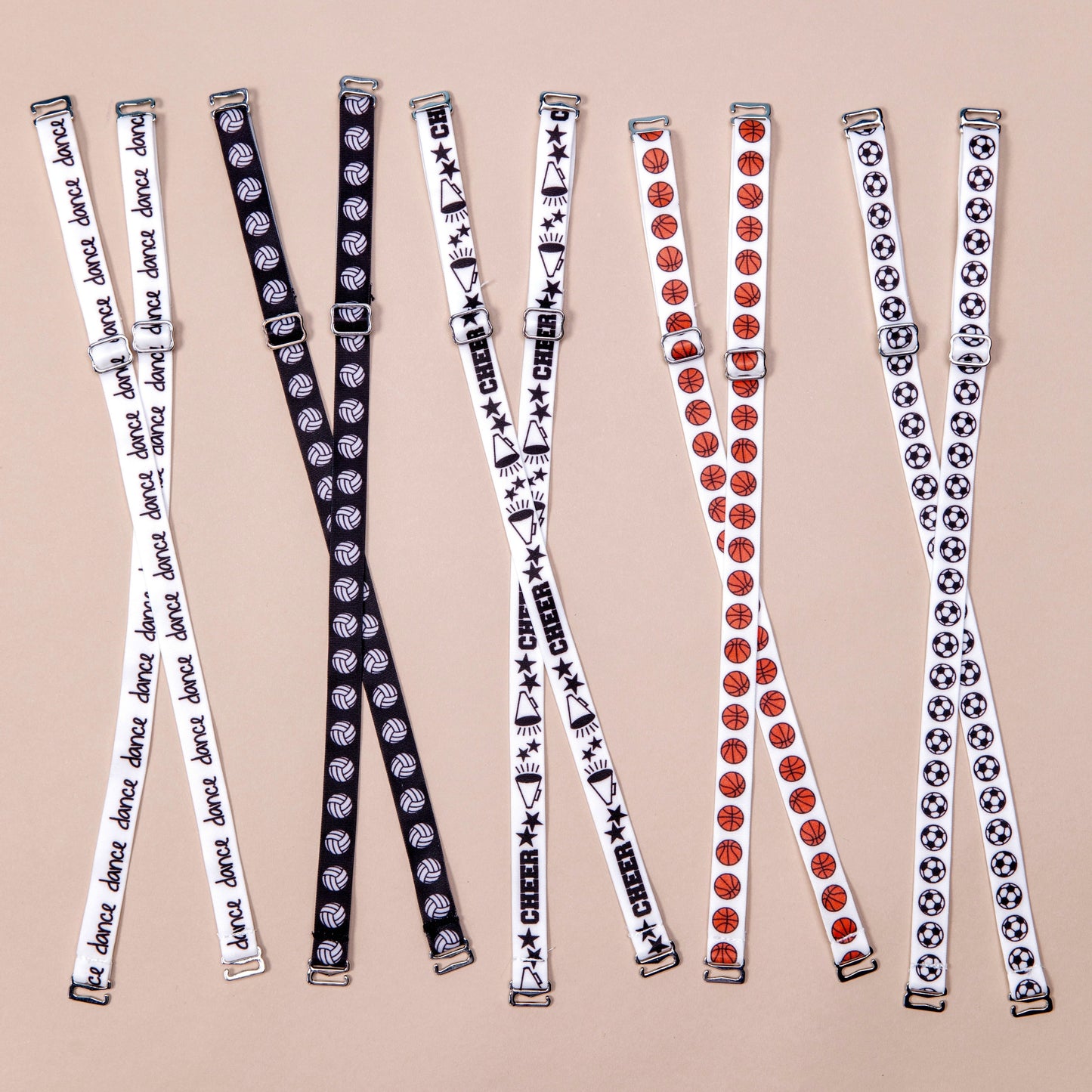 Bra straps laid out in criss cross formation from left to right: White straps with the word dance in black handwritten font repeating along length of material, Black straps with white volleyballs repeating along length of material, White straps with megaphones, black stars and the word Cheer in black block letters, alternating along length of material, white straps with orange basketballs repeating along length of material, white straps with black and white soccer repeating along length of material.