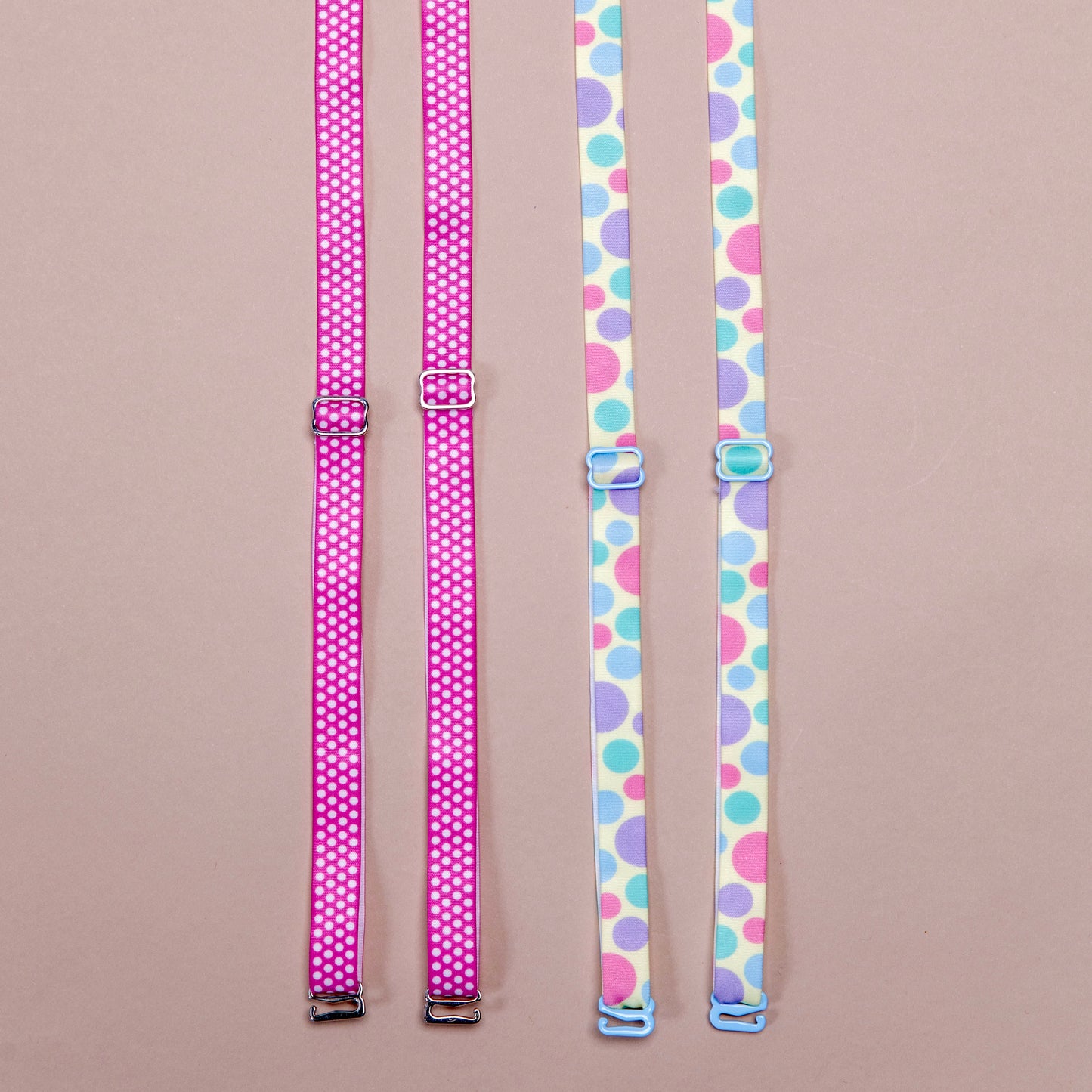 2 sets of interchangeable her-rah bra straps in polka dot patterns. Left: Strawberry Polka Her-Rah Straps - Dark pink strap with small uniform light pink polka dots repeating the length of the strap, featuring metal hardwear. Right: Spring Fling Her-Rah Straps - White straps with a mix of various size polka dots in light blue, periwinkle, and hot pink, featuring blue coated hardware.