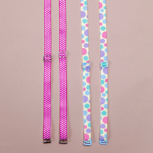 2 sets of interchangeable her-rah bra straps in polka dot patterns. Left: Strawberry Polka Her-Rah Straps - Dark pink strap with small uniform light pink polka dots repeating the length of the strap, featuring metal hardwear. Right: Spring Fling Her-Rah Straps - White straps with a mix of various size polka dots in light blue, periwinkle, and hot pink, featuring blue coated hardware.