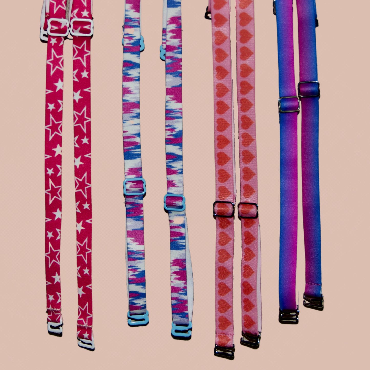 4 sets of Her-Rah interchangeable bra straps. From Left to right: Twinkle Her-Rah Straps - Hot Pink straps with a mix of solid white and outlined stars in varied sizes, featuring white coated hardware. 3-D Static - White straps with fading static/sound wave shapes in hot pink, gray, and blue, featuring blue coated hardware. Hearts - pink straps with red hearts repeating the length of the strap, featuring silver metal hardware.