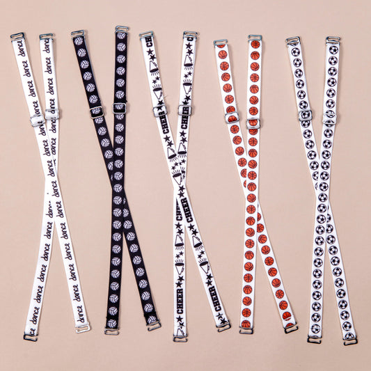 Bra straps laid out in criss cross formation from left to right: White straps with the word dance in black handwritten font repeating along length of material, Black straps with white volleyballs repeating along length of material, White straps with megaphones, black stars and the word Cheer in black block letters, alternating along length of material, white straps with orange basketballs repeating along length of material, white straps with black and white soccer repeating along length of material.